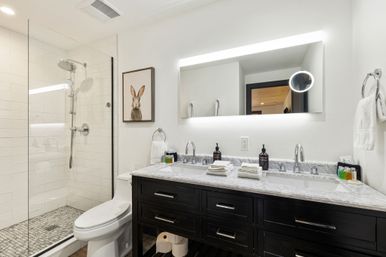 Spa-like modern bathroom with glass-enclosed walk-in shower and white subway tile, marble double vanity with backlit mirror and dark wood cabinets