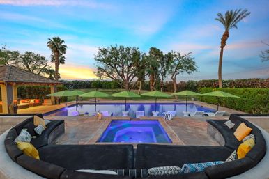 Sunset luxury backyard pool and illuminated spa with in-pool loungers under green umbrellas, curved outdoor sofa with colorful pillows, gazebo, palm trees and manicured lawn