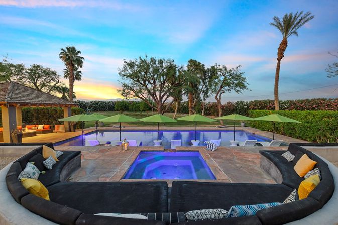 Sunset luxury backyard pool and illuminated spa with in-pool loungers under green umbrellas, curved outdoor sofa with colorful pillows, gazebo, palm trees and manicured lawn