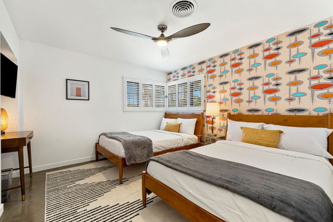 Bright mid-century modern bedroom with two wooden beds dressed in white linens and gray throws, playful colorful retro wallpaper accent wall, bedside lamp, ceiling fan, shuttered window and striped area rug