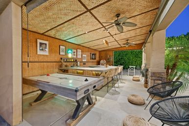 Tropical-style backyard covered patio and outdoor bar with an air hockey table, stone countertop and rattan barstools, woven bamboo ceiling with ceiling fans, string lights and lush greenery at dusk.