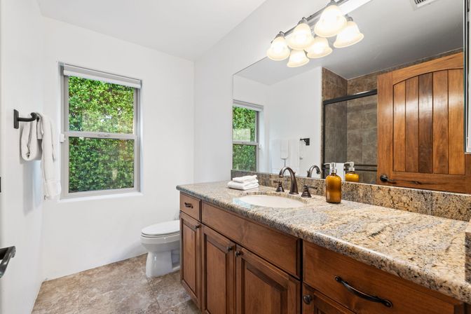 Bright cozy bathroom with granite countertop and undermount sink, wooden vanity with bronze faucet, tiled floor, glass shower, arched wood door, white towels and a window showing a green hedge.