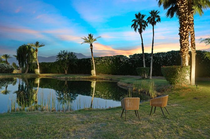 Two wicker chairs beside a palm-lined reflecting pond at sunset, pastel sky and distant mountains creating a serene desert-oasis scene