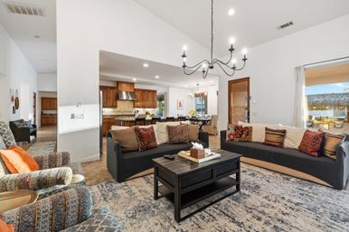 Sunlit open-concept living room and kitchen in a coastal waterfront home with vaulted ceiling and black chandelier, two sofas and patterned armchairs around a dark wood coffee table on a blue-gray rug, wooden kitchen cabinets with island, dining area, and sliding doors opening to a thatched-roof patio and water view.