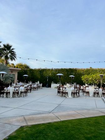 Outdoor patio reception setup with round tables draped in white, wooden cross-back chairs, string lights overhead, patio heaters, a palm tree and hedged garden under a clear blue sky