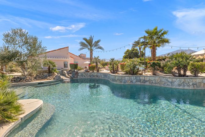 Sparkling lagoon-style backyard pool with stone edging, palm trees, desert landscaping, string lights and distant mountain views under a bright blue sky