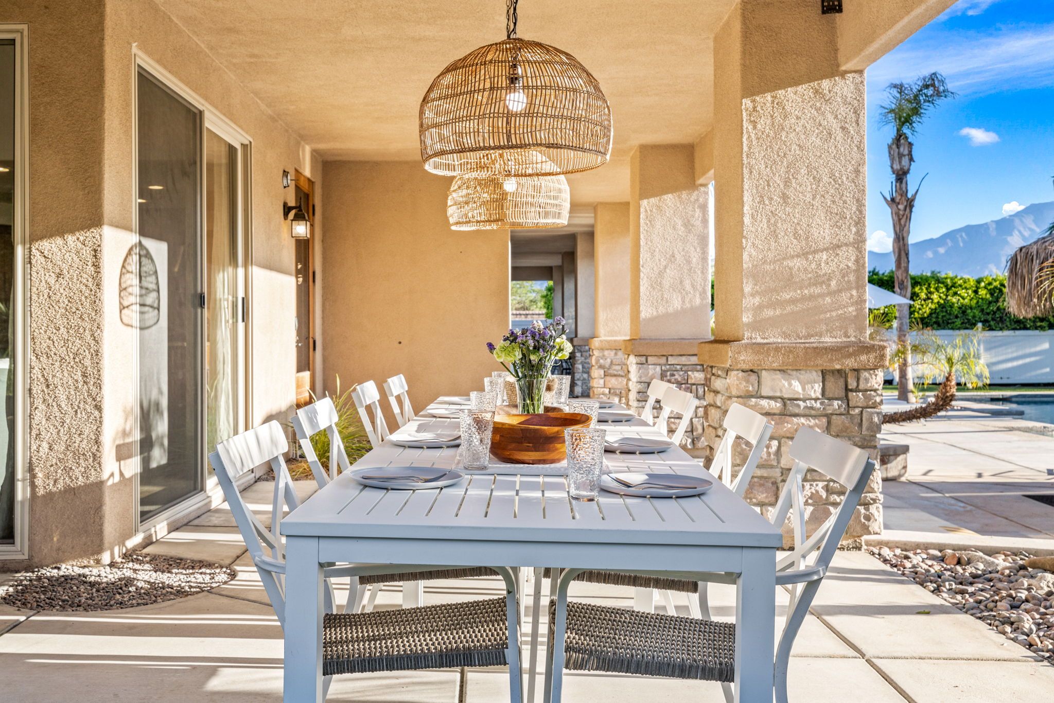 Sunlit covered backyard patio with a long white dining table set for eight, woven pendant lights overhead, a flower centerpiece, stone columns and poolside view with a palm tree and distant mountains.