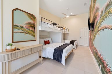 Tropical coastal bedroom with white bunk and twin beds, large palm-frond mural and framed palm art, light wood console, ceiling fan and wall-mounted TV — airy vacation-rental vibe.