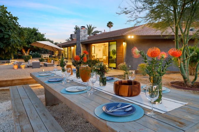 Sunset backyard patio with a long wooden picnic table set for dinner — blue place mats, wine glasses, orange flowers, gravel courtyard and modern single-story house in the background.