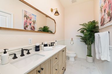Bright coastal-inspired powder room with light-wood double vanity, white quartz countertop, matte black faucets, gold-framed mirror, potted palm, pink building wall art and marble tile floor.
