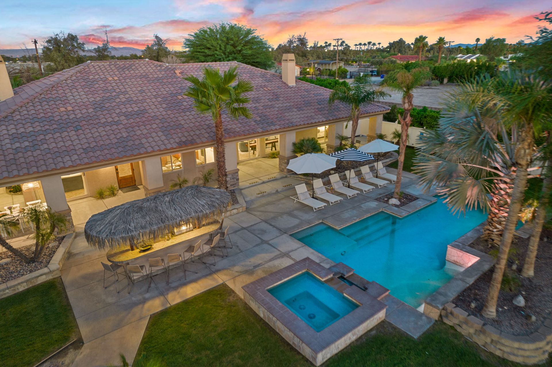 Sunset backyard oasis with a turquoise swimming pool and raised hot tub, tiled-roof house, thatched tiki bar with stools, row of chaise lounges and umbrellas, and palm trees