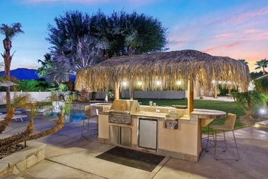 Tiki-style outdoor kitchen and bar with stainless grill beside a pool, palm trees and mountain sunset