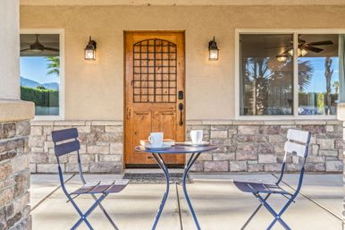 Sunlit front porch with a wooden arched door, stone veneer facade, two wall lanterns and a small round bistro table set with two cups and folding chairs.