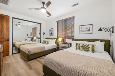 Bright modern bedroom interior with two olive-green upholstered beds, beige knit blankets and fern-print pillows, wooden blinds, mirrored closet doors, ceiling fan, bedside lamp and warm wood floors.