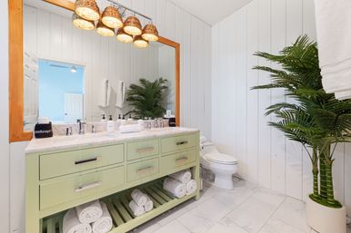 Bright spa-like coastal bathroom with sage green double-sink vanity, marble countertop, wicker pendant lights, large wood-framed mirror, rolled towels, potted palm, and white shiplap walls