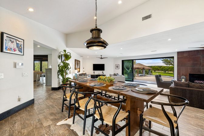 Open-concept dining and living room with rustic wooden high-top table and woven black chairs under an industrial pendant, sliding glass doors opening to a patio gazebo and vineyard views