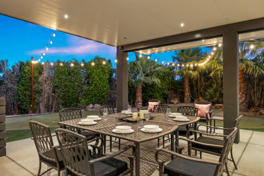 Covered backyard patio dining area at dusk with a metal table set for six, plates and candles, string lights overhead and palm trees and hedges in a landscaped yard.