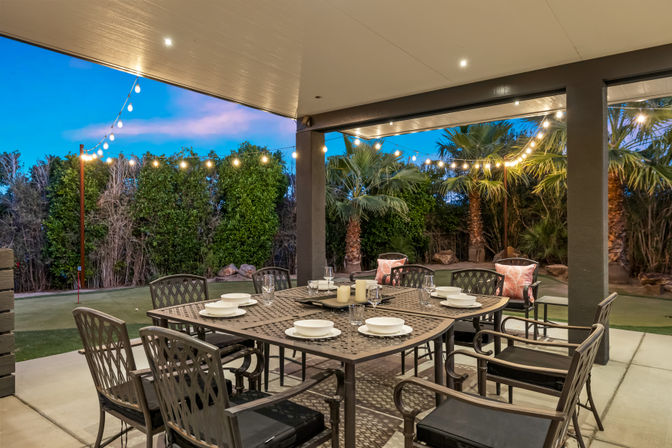 Covered backyard patio dining area at dusk with a metal table set for six, plates and candles, string lights overhead and palm trees and hedges in a landscaped yard.