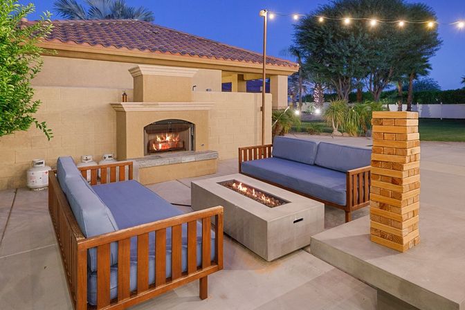 Cozy evening backyard patio with a lit built-in fireplace, modern concrete fire pit between wooden lounge sofas with blue cushions, string lights overhead and palm trees in the background