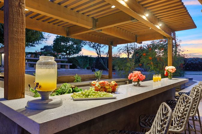Outdoor patio bar at sunset under a wooden pergola with a marble countertop, glass beverage dispenser, plate of grapes, basket of apples, pink flower vases and woven bar stools.