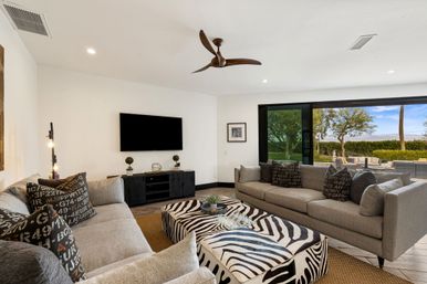 Bright modern living room with gray sofas, zebra-print ottoman, wooden ceiling fan and wall-mounted TV, sliding glass doors opening to patio and landscaped backyard with palm tree