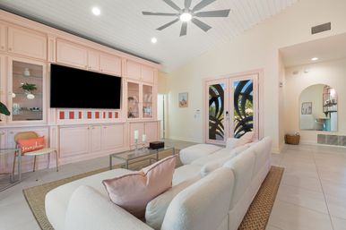 Bright modern living room featuring pastel pink built-in cabinetry with a large wall-mounted TV, plush white sectional on a woven rug, glass coffee table, ceiling fan, and decorative glass double doors opening to a sunny garden.