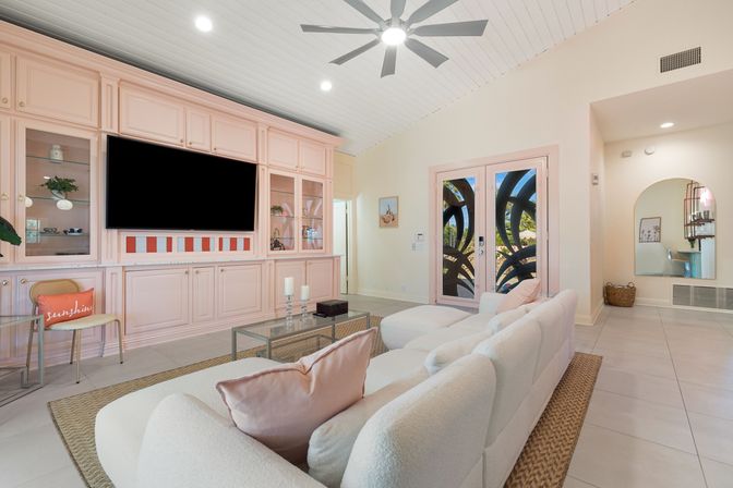 Bright modern living room featuring pastel pink built-in cabinetry with a large wall-mounted TV, plush white sectional on a woven rug, glass coffee table, ceiling fan, and decorative glass double doors opening to a sunny garden.