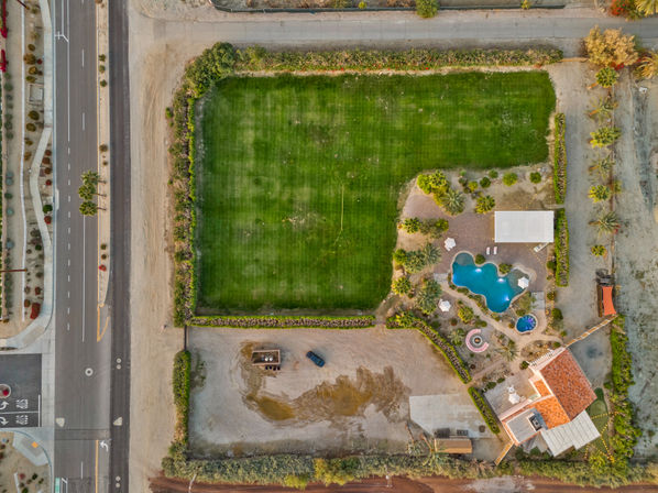 Aerial drone view of a desert estate with a large rectangular green lawn, kidney-shaped blue swimming pool, palm trees, patio area and adjacent roadway