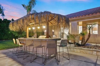 Inviting suburban backyard tiki-style outdoor bar and kitchen with rattan barstools, thatched palapa roof, hanging globe lights and palm trees at dusk