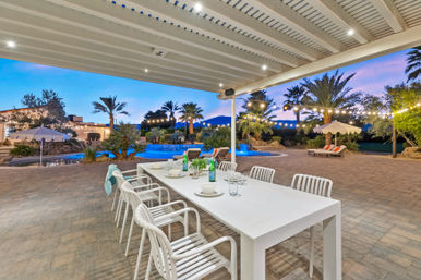 Covered backyard patio with white outdoor dining table and chairs on paver patio, resort-style pool, palm trees, string lights and mountain sunset view