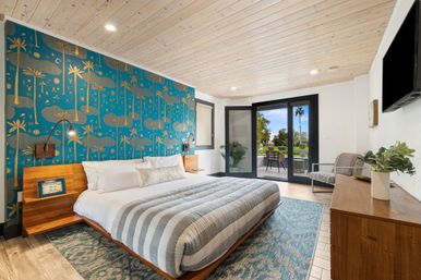 Modern coastal king bedroom with teal palm-patterned accent wall, wood-plank ceiling, striped bedding and sliding patio doors opening to palm-lined outdoor seating.