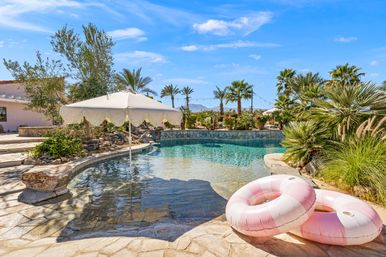 Sunlit resort-style backyard pool with a scalloped white umbrella over a shallow entry, two pink striped inflatable rings on the stone deck, palm trees and desert hills under a bright blue sky.