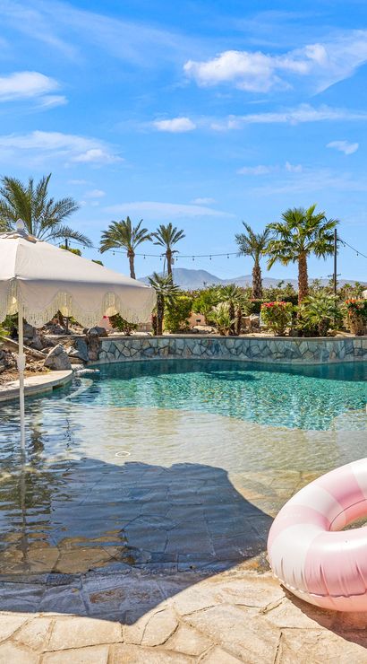 Sunlit resort-style backyard pool with a scalloped white umbrella over a shallow entry, two pink striped inflatable rings on the stone deck, palm trees and desert hills under a bright blue sky.