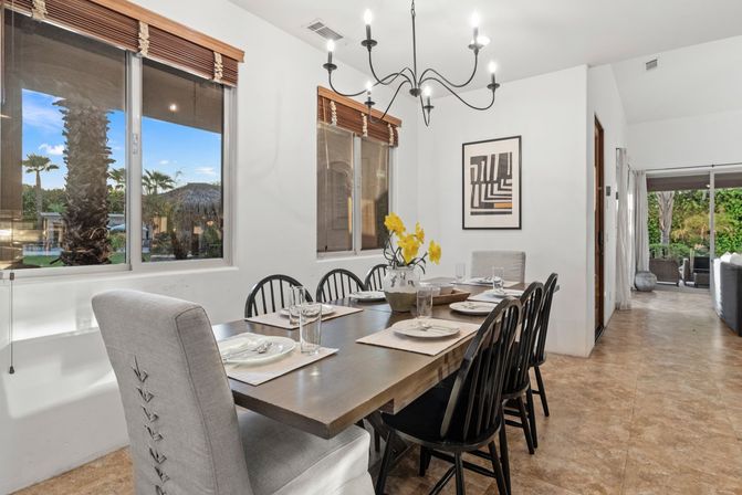 Bright modern dining room with a wooden table set for six, black spindle chairs and upholstered end chairs, black chandelier overhead, yellow floral centerpiece, and large windows framing palm trees and a pool in a tropical backyard.