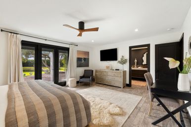 Sunlit modern master bedroom with beige-gray striped quilt, wooden ceiling fan, black-framed glass doors opening to a garden lawn, rustic wood dresser under a wall-mounted TV, cozy armchair, desk with vase, and neutral airy decor.