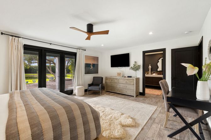 Sunlit modern master bedroom with beige-gray striped quilt, wooden ceiling fan, black-framed glass doors opening to a garden lawn, rustic wood dresser under a wall-mounted TV, cozy armchair, desk with vase, and neutral airy decor.