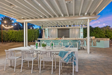 Covered outdoor patio dining area with long white table and chairs, turquoise bar with stools, built-in stainless steel grill, string lights and potted plants on a paved stone floor at dusk