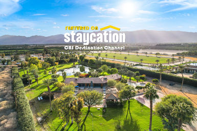 Aerial view of a lush desert oasis estate with palm trees, green lawns, a reflective pond, tiled-roof house and distant mountains under a bright blue sky