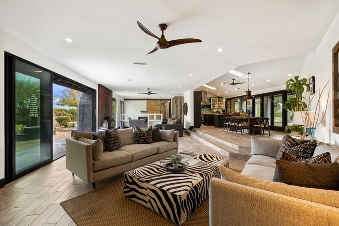 Open-concept modern living room and kitchen with sliding glass doors to a sunlit patio, neutral sofas and accent pillows, zebra-print ottoman, wood-look floors, ceiling fans, pendant lights and a raised dining area.