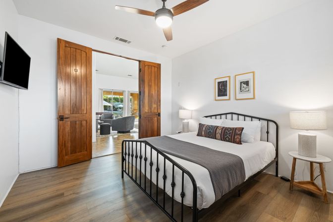 Bright, airy bedroom with black metal bed dressed in white linens and a patterned bolster, wood-plank floors, ceiling fan, twin bedside lamps, and warm wooden double doors opening to a sunlit living area and outdoor patio with umbrella and greenery.