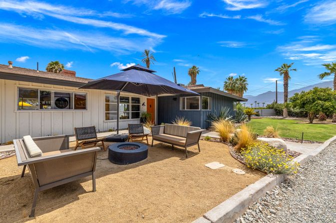 Sunny desert backyard patio with midcentury modern seating around a round fire pit under a black umbrella, palm trees, drought-tolerant landscaping and mountain views