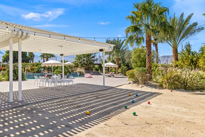 Sunny desert poolside patio with white pergola casting striped shadows over an outdoor dining set, sandy bocce court with colorful balls, palm trees, umbrellas and mountains in the background — relaxed Southern California desert oasis.