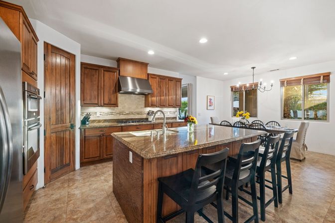 Open-plan family kitchen with large granite island and breakfast bar, rich wood cabinets, stainless steel appliances, tile floor, and sunny dining nook with chandelier