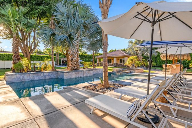 Sun-drenched backyard pool framed by palm trees, white lounge chairs and umbrellas beside a stone deck and outdoor cabana