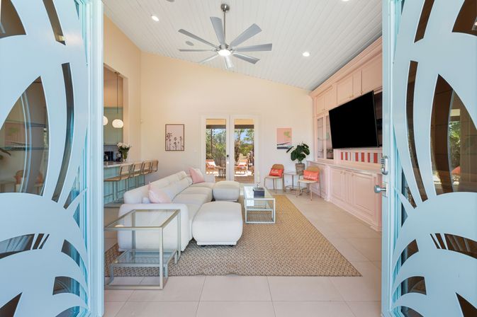 Sunny coastal-style living room framed by turquoise decorative entry doors, featuring a white sectional on a woven rug, glass coffee tables, pastel-pink built-in media wall with TV, bar stools, ceiling fan, and French doors opening to a patio.