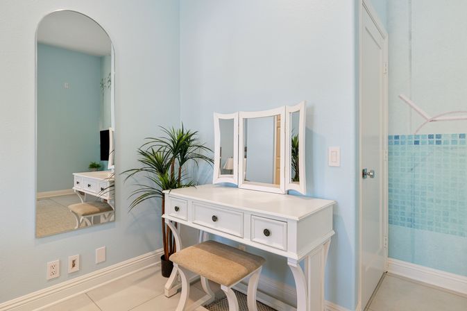 White vintage-style vanity with tri-fold mirror and cushioned stool against a pale blue wall, arched wall mirror and potted palm creating a bright coastal dressing nook