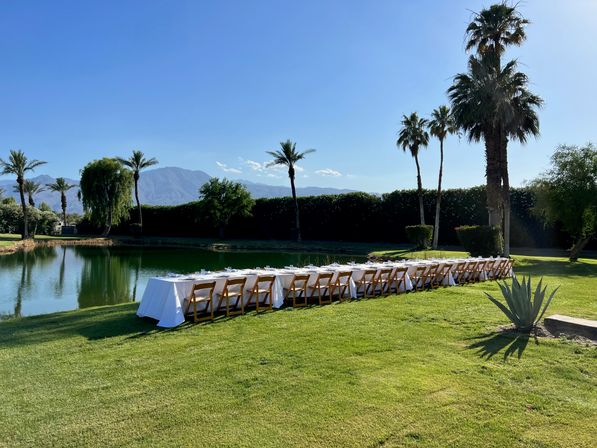Long al fresco banquet table with white tablecloths and wooden chairs on a green lawn beside a calm lake, framed by palm trees and distant mountains under a clear blue sky.