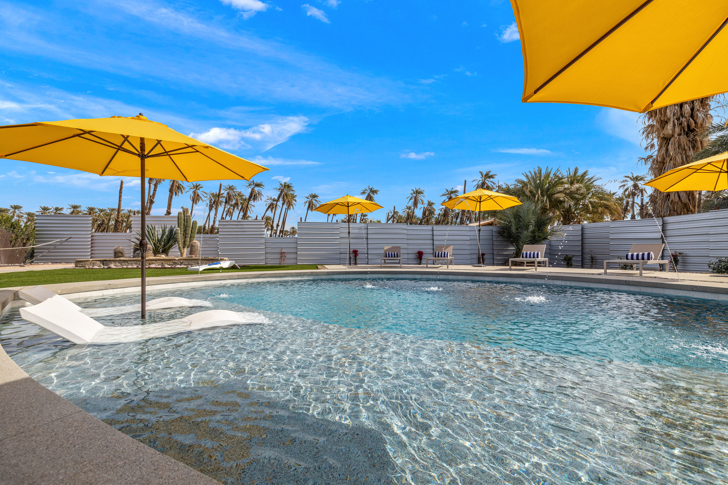 Resort-style California desert pool with bright yellow umbrellas, in-water loungers on a shallow tanning ledge, striped chaise lounges and palm trees under a vivid blue sky.
