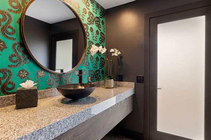 Modern powder room with bold teal snake-print wallpaper, round mirror, black vessel sink, granite countertop, white orchid and frosted glass door.