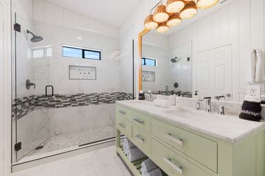 Bright modern bathroom with frameless glass walk-in shower, marble tile and gray mosaic accent band, mint-green double vanity with marble countertop, woven pendant lights, large mirror and neatly rolled towels.